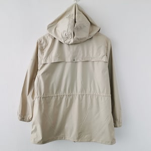 May include: A beige hooded jacket featuring long sleeves with elastic cuffs, a drawstring waist, and a horizontal back yoke flap with a single snap button closure. The lightweight garment is shown from the back, displaying a smooth fabric texture.