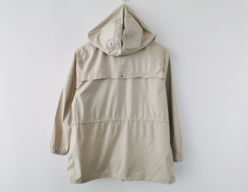 May include: A beige hooded jacket featuring long sleeves with elastic cuffs, a drawstring waist, and a horizontal back yoke flap with a single snap button closure. The lightweight garment is shown from the back, displaying a smooth fabric texture.