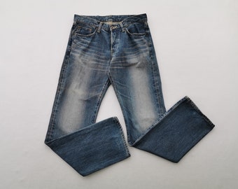 Puede incluir: Unos pantalones vaqueros de mezclilla azul con un lavado desgastado en los muslos, cierre de botones, cinco bolsillos y corte de pierna recta. El tejido presenta detalles de desgaste visibles en las caderas y un color azul medio.