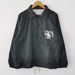 May include: Black coach jacket with a button-up front and a collar. The jacket features a white graphic of a devil with a pitchfork on the front. The jacket is hanging on a wooden hanger against a white background.