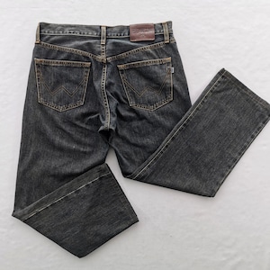 Può includere: Jeans in denim grigio scuro con un classico design a cinque tasche. I jeans presentano una toppa in pelle marrone sul retro e il logo del marchio sulle tasche posteriori. I jeans sono stesi su una superficie bianca.