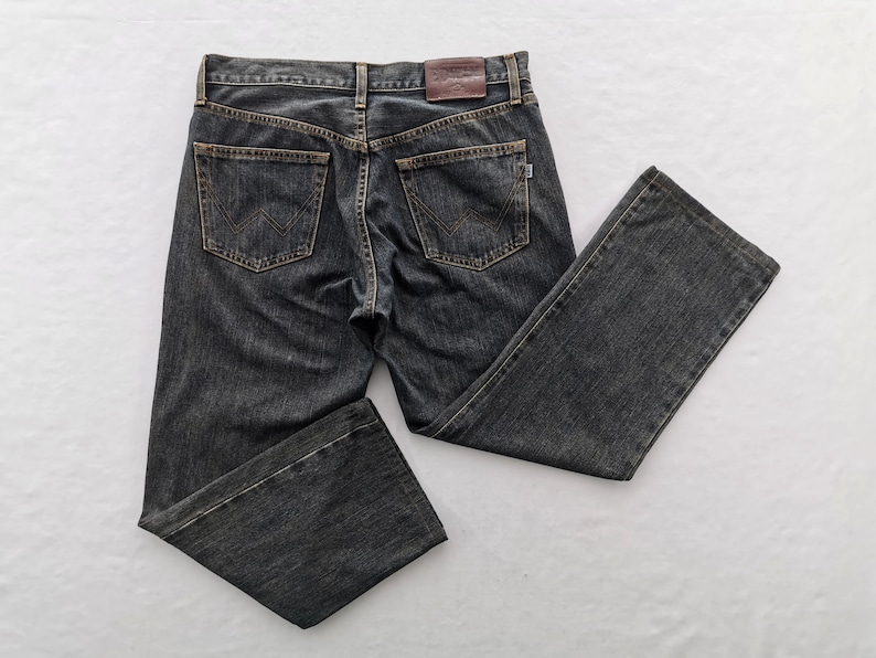 May include: Dark grey denim jeans with a classic five-pocket design. The jeans feature a brown leather patch on the back and the brand's logo on the back pockets. The jeans are laid flat on a white surface.