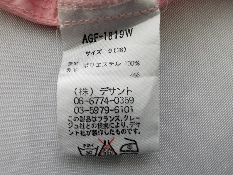 May include: A white fabric clothing care label with black text, featuring the code AGF-1819W, size 9 (38), and 100% polyester composition. It includes Japanese text, contact phone numbers, and three laundry instruction symbols at the bottom.
