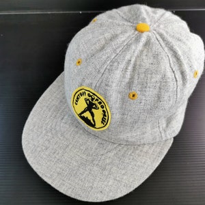 Könnte beinhalten: Graue Baseballkappe mit gebogenem Schirm. Auf der Kappe befindet sich ein gelber Aufnäher mit dem Text "CENTRAL OOPS PROGRESS" und einer Personengrafik. Gelbe Nähte und ein gelber Knopf runden das Design ab.