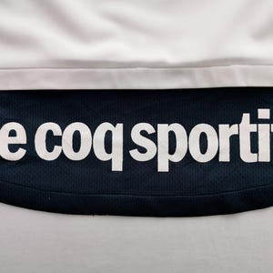 K&ouml;nnte beinhalten: Nahaufnahme eines wei&szlig;en und marineblauen Sportkleidungsst&uuml;cks. Der Text "le coq sportif" ist in wei&szlig;en Blockbuchstaben auf dem marineblauen Stoff zu sehen. Der obere Teil ist wei&szlig;.