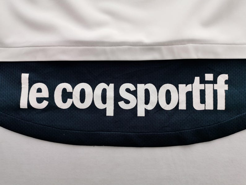 K&ouml;nnte beinhalten: Nahaufnahme eines wei&szlig;en und marineblauen Sportkleidungsst&uuml;cks. Der Text "le coq sportif" ist in wei&szlig;en Blockbuchstaben auf dem marineblauen Stoff zu sehen. Der obere Teil ist wei&szlig;.