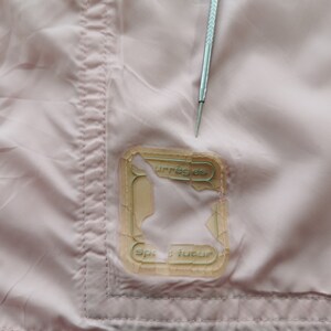 May include: A light pink fabric garment featuring a rectangular, semi-transparent patch with the text "courr&egrave;ges" and "sport futur" printed on it. A silver metal tool with a pointed tip is positioned vertically above the patch.