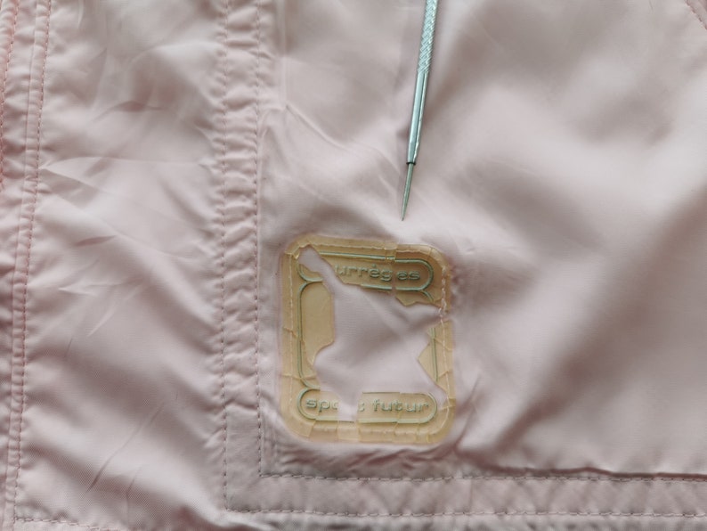 May include: A light pink fabric garment featuring a rectangular, semi-transparent patch with the text "courr&egrave;ges" and "sport futur" printed on it. A silver metal tool with a pointed tip is positioned vertically above the patch.