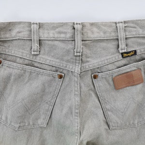 May include: Close-up of the back of light gray Wrangler jeans. Features include two back pockets, a leather patch, and the Wrangler logo. The jeans have belt loops and copper-colored rivets.