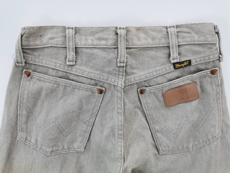 May include: Close-up of the back of light gray Wrangler jeans. Features include two back pockets, a leather patch, and the Wrangler logo. The jeans have belt loops and copper-colored rivets.