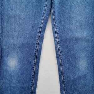 May include: Close-up of blue denim jeans. The jeans have a classic five-pocket design, with visible stitching and a faded appearance. The fabric shows signs of wear, adding to the vintage aesthetic. The jeans are a medium wash, suitable for casual wear.