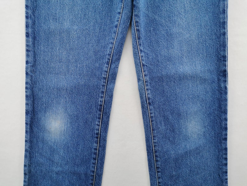 May include: Close-up of blue denim jeans. The jeans have a classic five-pocket design, with visible stitching and a faded appearance. The fabric shows signs of wear, adding to the vintage aesthetic. The jeans are a medium wash, suitable for casual wear.