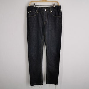 May include: Dark wash denim jeans with a classic five-pocket design, featuring yellow stitching and a button fly. The jeans are hanging on a wooden hanger against a plain white wall.