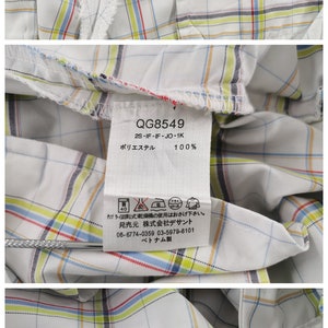 Op de afbeelding: Een wit golf shirt met een rood, blauw en geel ruitpatroon. Het shirt heeft een label met de tekst "lecoqsportif GOLF COLLECTION" en "QG8549 2S-IF-IF-JO-1K" in het Japans. Het label bevat ook wasinstructies en de tekst "100% ポリエステル" in het Japans.