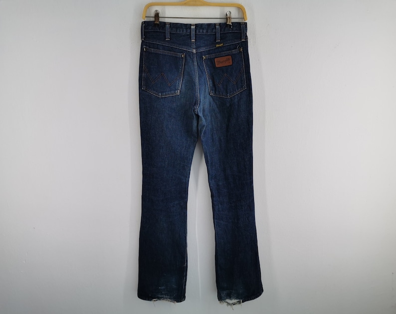 Peut inclure: Jean bleu fonc&eacute; avec une conception classique &agrave; cinq poches, suspendu &agrave; un cintre en bois. Le jean pr&eacute;sente un patch en cuir marron avec le logo "Wrangler" sur la poche arri&egrave;re. L'ourlet inf&eacute;rieur montre une certaine usure.