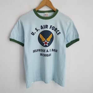 May include: Light blue ringer t-shirt with dark green trim and the text "U.S. AIR FORCE SELFRIDGE A.F. BASE MICHIGAN" in navy blue. The shirt features a navy blue emblem with gold wings and a red star.