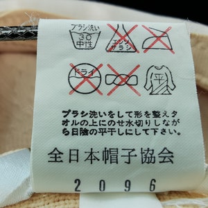 Puede incluir: Primer plano de una etiqueta con instrucciones de cuidado y texto en japon&eacute;s. La etiqueta incluye s&iacute;mbolos de lavado, secado y planchado. El texto dice "全日本帽子協会" y "2096". La etiqueta est&aacute; adherida a una tela beige.