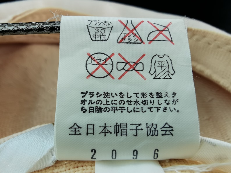 Puede incluir: Primer plano de una etiqueta con instrucciones de cuidado y texto en japon&eacute;s. La etiqueta incluye s&iacute;mbolos de lavado, secado y planchado. El texto dice "全日本帽子協会" y "2096". La etiqueta est&aacute; adherida a una tela beige.
