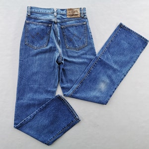 May include: Blue denim jeans with a classic five-pocket design. The jeans feature a faded wash and a leather patch with the brand name. The back pockets have a signature embroidered design. The jeans are laid flat on a white surface.