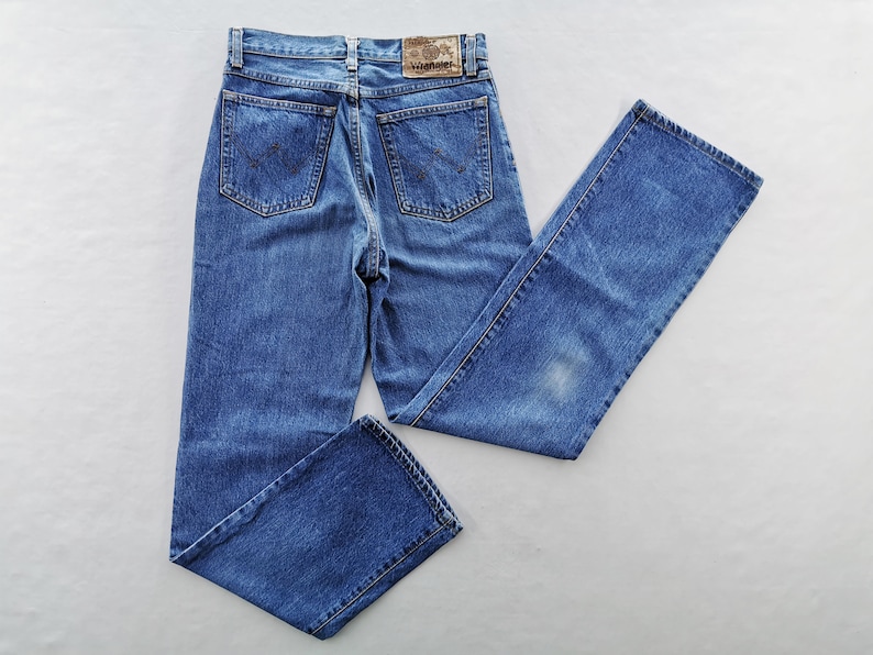May include: Blue denim jeans with a classic five-pocket design. The jeans feature a faded wash and a leather patch with the brand name. The back pockets have a signature embroidered design. The jeans are laid flat on a white surface.