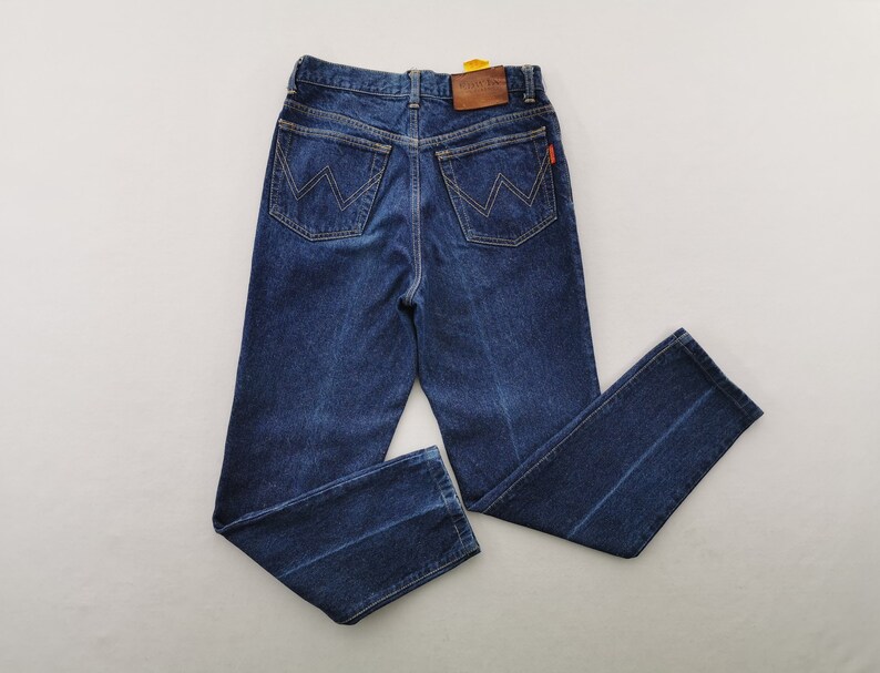 May include: Dark blue denim jeans with a classic five-pocket design. Features a leather patch and a red tag. The back pockets have a distinctive 'W' stitching. The jeans are laid flat, showcasing the straight leg cut.
