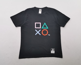 Puede incluir: Una camiseta negra de manga corta con los símbolos del mando de PlayStation en rosa, verde, azul y naranja en el pecho. Un pequeño parche blanco con el logotipo de PlayStation Official Licensed Product se encuentra en la esquina inferior derecha.