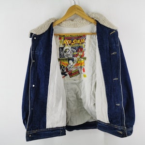 May include: A dark blue denim jacket featuring a white sherpa collar and a white interior lining. The back interior panel displays a colorful Red Sonja Marvel Comics graphic print with yellow and red text, including the title Red Sonja.