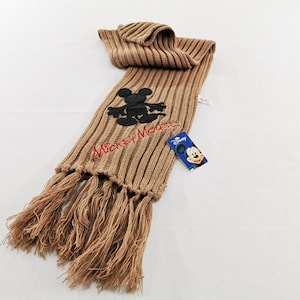 Peut inclure: Une écharpe en tricot côtelé beige avec une silhouette noire brodée de Mickey Mouse et le texte Mickey Mouse cousu en rouge. L'écharpe présente de longues franges aux extrémités et une étiquette de marque Disney bleue attachée sur le côté.