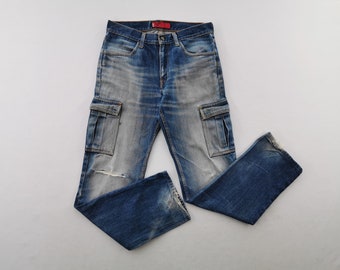 Puede incluir: Pantalones cargo de mezclilla azul con un lavado desgastado, dos bolsillos laterales con solapa, cierre de botones y una pequeña etiqueta roja de marca en la cintura. La tela muestra detalles desgastados, incluyendo un pequeño roto en la rodilla izquierda.