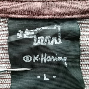 Pode incluir: Close-up de uma etiqueta preta com texto branco e um design gr&aacute;fico. A etiqueta diz "&copy; K. Haring" e "L". A etiqueta est&aacute; presa a um tecido listrado marrom e branco.