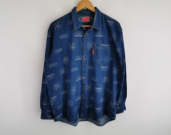 Puede incluir: Una camisa de manga larga en mezclilla azul con un patrón estampado en blanco de logotipos circulares y el texto Drug Store. La camisa incluye un bolsillo en el pecho con una pequeña etiqueta roja y una etiqueta roja en el cuello.