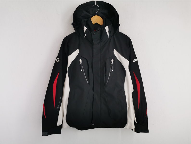 Pode incluir: Casaco preto e branco com capuz e detalhes vermelhos. O casaco tem fecho de correr, v&aacute;rios bolsos e a palavra "GO" na manga. O casaco est&aacute; pendurado num cabide de madeira.