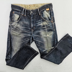 May include: Dark wash denim jeans with distressed details. The jeans have a classic five-pocket design, a button and zip closure, and a faded, worn appearance. The jeans are folded on a white surface.