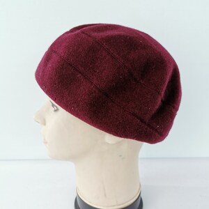 May include: A burgundy-colored, wool beret-style hat. The hat has a rounded crown with vertical stitched details. The hat is displayed on a mannequin head against a white background. The hat is a solid color.