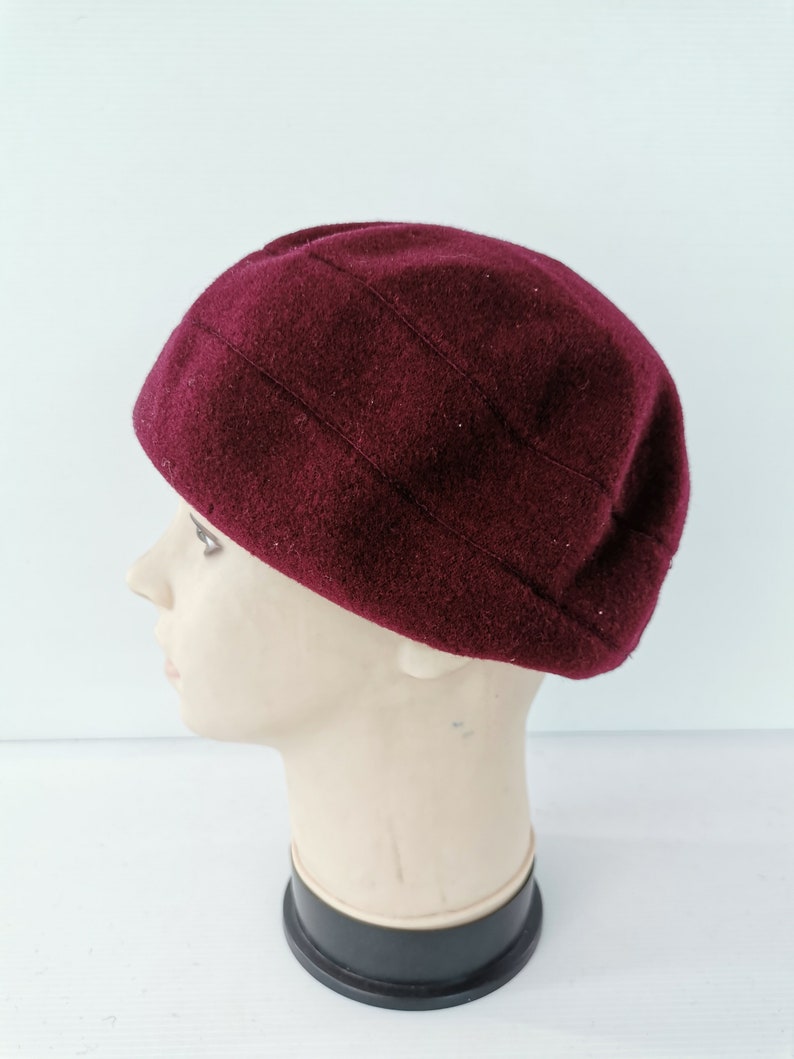 May include: A burgundy-colored, wool beret-style hat. The hat has a rounded crown with vertical stitched details. The hat is displayed on a mannequin head against a white background. The hat is a solid color.