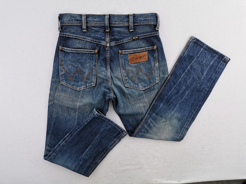 Może przedstawiać: Niebieskie dżinsy Wrangler z klasycznym pięciokieszeniowym designem. Dżinsy mają logo Wrangler na tylnej kieszeni i zużyty wygląd. Dżinsy są ułożone płasko na białej powierzchni.