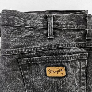 May include: Close-up of black denim jeans with a faded wash. The jeans feature a back pocket with a brown leather Wrangler label. The waistband has belt loops and a button closure. The denim has a textured appearance.
