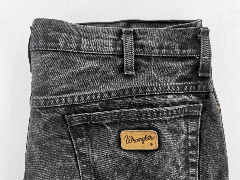 May include: Close-up of black denim jeans with a faded wash. The jeans feature a back pocket with a brown leather Wrangler label. The waistband has belt loops and a button closure. The denim has a textured appearance.