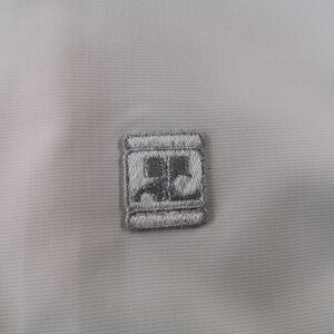 Puede incluir: Primer plano de una tela gris claro con un peque&ntilde;o parche bordado. El parche es de forma cuadrada con un borde plateado y un dise&ntilde;o gris en el interior. La tela tiene un patr&oacute;n sutil y texturizado.