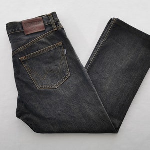 May include: Dark grey denim jeans with a classic five-pocket design. Features a brown leather patch and a back pocket with a stitched logo. The jeans are folded, showcasing the straight leg cut and the worn look of the fabric.