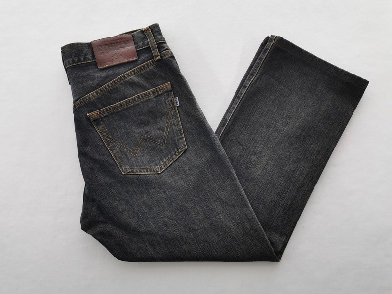 May include: Dark grey denim jeans with a classic five-pocket design. Features a brown leather patch and a back pocket with a stitched logo. The jeans are folded, showcasing the straight leg cut and the worn look of the fabric.