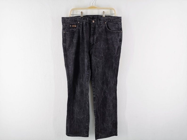 May include: Black denim jeans with a faded, textured appearance. The jeans have a classic five-pocket design, a button and zipper closure, and are hanging on a metal hanger against a white background. The jeans are a size 40.