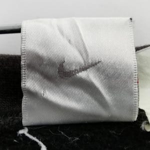 K&ouml;nnte beinhalten: Nahaufnahme eines wei&szlig;en Stoffetiketts mit dem grauen Nike-Logo. Das Etikett ist rechteckig mit einem gen&auml;hten Rand. Der Hintergrund ist dunkel und unscharf.