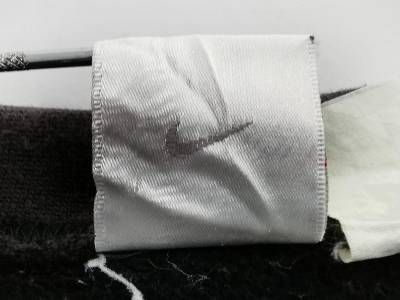 K&ouml;nnte beinhalten: Nahaufnahme eines wei&szlig;en Stoffetiketts mit dem grauen Nike-Logo. Das Etikett ist rechteckig mit einem gen&auml;hten Rand. Der Hintergrund ist dunkel und unscharf.