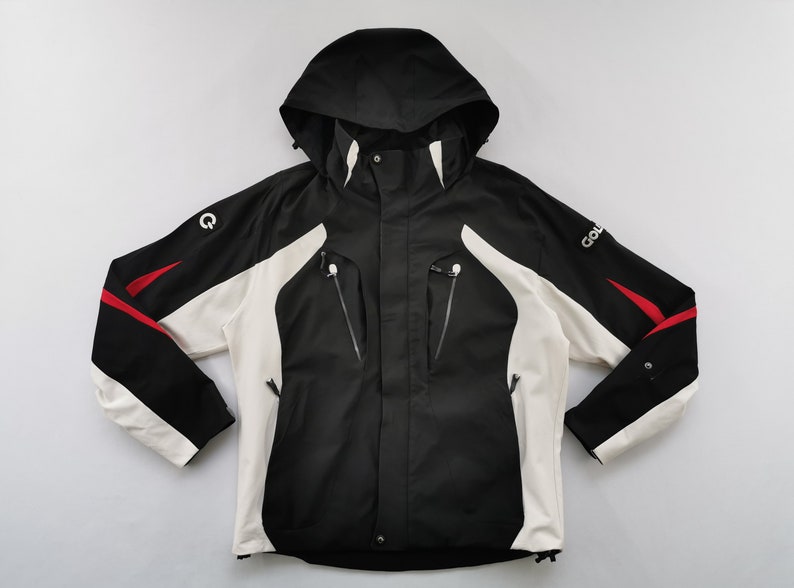 Pode incluir: Casaco com capuz preto, branco e vermelho com fecho de correr e v&aacute;rios bolsos. O casaco tem capuz e mangas pretas com pain&eacute;is brancos na frente e detalhes em vermelho. A palavra "GOL" &eacute; vis&iacute;vel na manga.