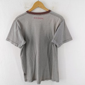 Pode incluir: Camiseta listrada cinza e branca com gola marrom e mangas curtas. A camisa tem um design costurado nos ombros e o texto "K. Haring" em vermelho. Est&aacute; pendurada em um cabide de madeira.