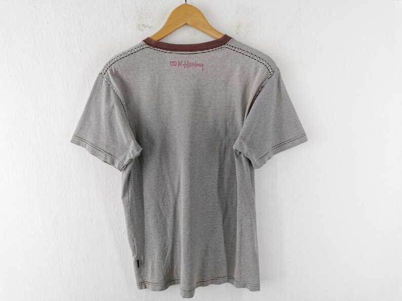 Pode incluir: Camiseta listrada cinza e branca com gola marrom e mangas curtas. A camisa tem um design costurado nos ombros e o texto "K. Haring" em vermelho. Est&aacute; pendurada em um cabide de madeira.