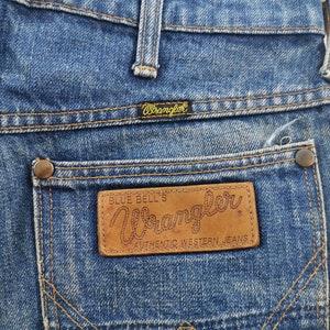 K&ouml;nnte beinhalten: Nahaufnahme einer blauen Wrangler-Jeans. Die Ges&auml;&szlig;tasche hat einen braunen Lederaufn&auml;her mit dem Wrangler-Logo und dem Text "Authentic Western Jeans". Die Jeans hat ein klassisches Design mit Kupfernieten und N&auml;hten.