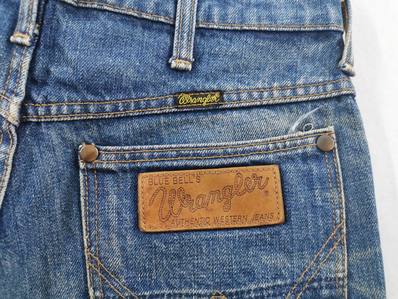 K&ouml;nnte beinhalten: Nahaufnahme einer blauen Wrangler-Jeans. Die Ges&auml;&szlig;tasche hat einen braunen Lederaufn&auml;her mit dem Wrangler-Logo und dem Text "Authentic Western Jeans". Die Jeans hat ein klassisches Design mit Kupfernieten und N&auml;hten.