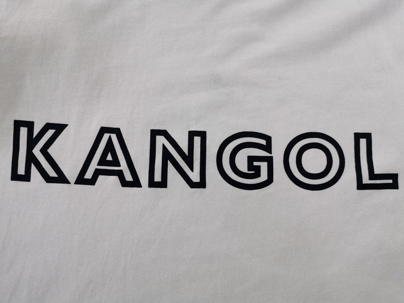 Peut inclure: Tissu blanc avec le mot "KANGOL" en noir, en gras et soulign&eacute;. Les lettres sont espac&eacute;es uniform&eacute;ment et centr&eacute;es, cr&eacute;ant un design &eacute;pur&eacute; et moderne. Le tissu semble lisse.